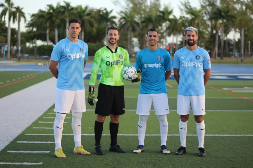 Four Dade County FC players in the club's home, goalkeeper and third kits on the turf at Traz Powell Stadium, palm trees behind them.