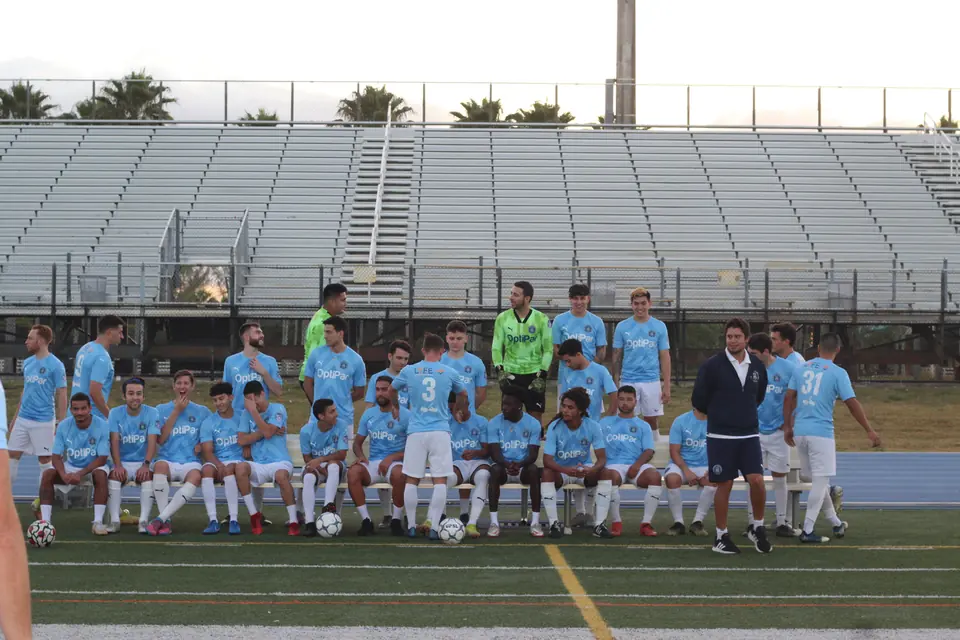 The Dade County FC first-team squad and coaching staff along the bench at their ground at dusk.