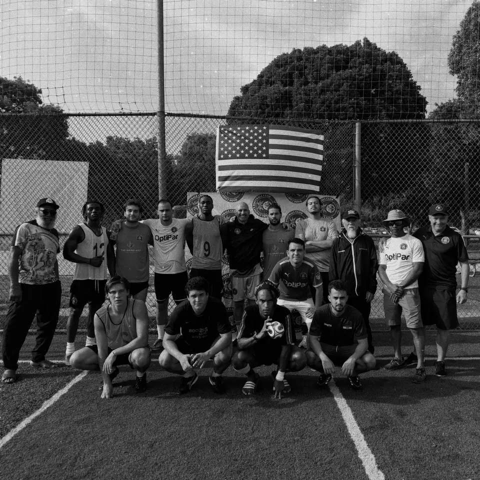 Around eighteen adult players and coaches in Dade County FC training tops after a community session, photographed in black and white in front of the club's banner.