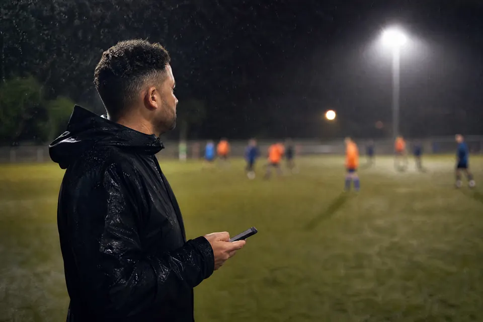 A league organizer in a rain jacket on a floodlit touchline, holding a phone while a game goes on behind