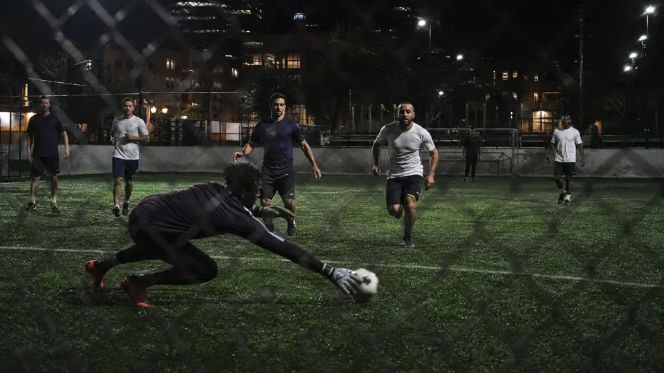 Night five-a-side soccer game on floodlit artificial turf, a goalkeeper diving for a low shot